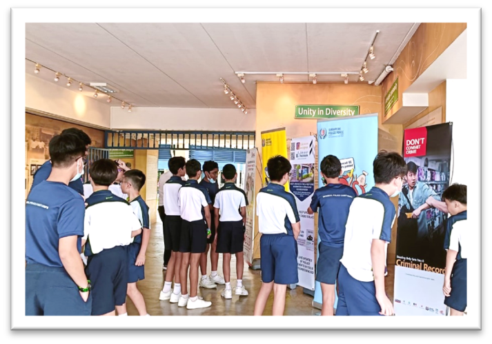 Students in school uniforms gather around informational banners including "Don't Commit Crime" and "Unity In Diversity" indoors.
